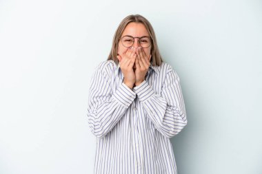 Mavi arka planda izole edilmiş genç beyaz kadın bir şeye gülüyor, elleriyle ağzını kapatıyor..