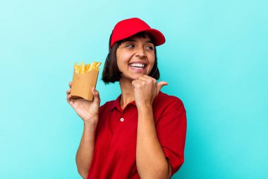 Genç melez kadın fast food restoranı çalışanı elinde kızartmalarla mavi arka planda izole edilmiş, baş parmak uzakta, gülen ve kaygısız..