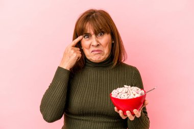 Middle age caucasian woman holding bowl of cereals isolated on pink background showing a disappointment gesture with forefinger.