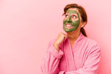 Middle age caucasian woman wearing a facial mask isolated on pink background looking sideways with doubtful and skeptical expression.