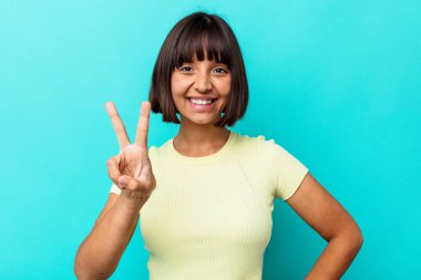 Mavi arka planda izole edilmiş genç melez kadın zafer işareti gösteriyor ve gülümsüyor..