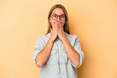Sarı arka planda izole edilmiş genç beyaz kadın elleriyle ağzını kapatırken şok oldu..