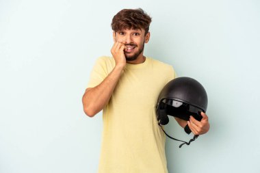 Mavi arka planda izole edilmiş, tırnak yiyen, gergin ve endişeli motosiklet kaskı tutan genç bir ırk..