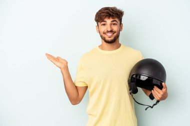 Mavi arka planda izole edilmiş motosiklet kaskı tutan melez genç bir adam bir avuç içinde fotokopi alanı ve belinde bir el daha gösteriyor..
