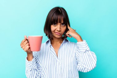 Elinde pembe kupa tutan genç bir ırk kadını mavi arka planda izole edilmiş, parmağıyla tapınağı işaret ediyor, düşünüyor, bir göreve odaklanıyor..