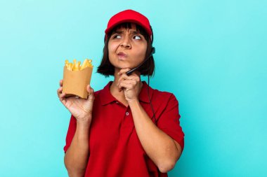 Genç melez kadın fast food restoranı çalışanı elinde patates kızartması ile mavi arka planda izole edilmiş şüpheli ve şüpheci bir ifadeyle yana bakıyor..