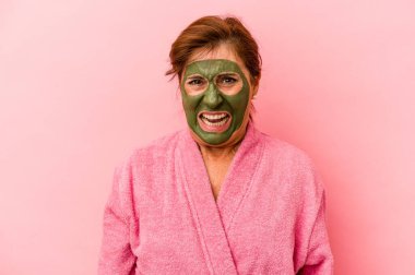 Middle age caucasian woman wearing a facial mask isolated on pink background screaming very angry and aggressive.