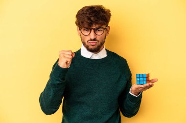Young mixed race man holding a Rubiks cube isolated on yellow background showing fist to camera, aggressive facial expression. 