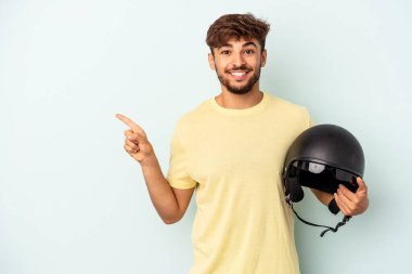 Mavi arka planda izole edilmiş motosiklet kaskı tutan genç melez adam gülümsüyor ve boş alanda bir şey gösteriyor..