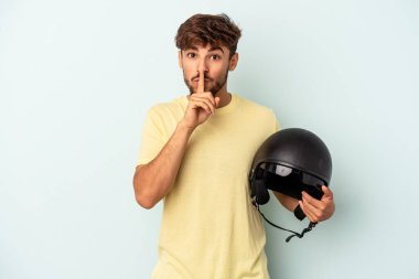 Mavi arka planda izole edilmiş motosiklet kaskı tutan genç melez bir adam ya sır saklıyor ya da sessizlik istiyor..