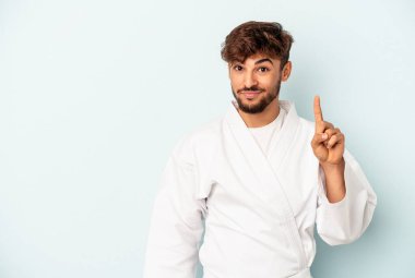 Genç melez karate yapıyor. Mavi arka planda izole edilmiş. Parmağıyla bir numarayı gösteriyor..