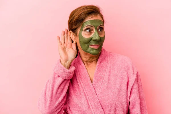 Middle age caucasian woman wearing a facial mask isolated on pink background trying to listening a gossip.