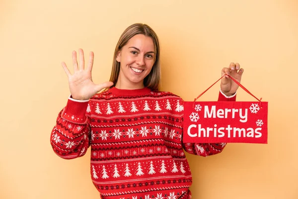 年轻的高加索女人拿着一块黄色背景的快乐的圣诞牌匾 笑容满面 用手指展示第五位 — 图库照片