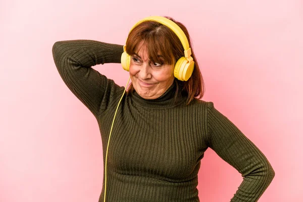 Middle age caucasian woman listening to music isolated on pink background touching back of head, thinking and making a choice.