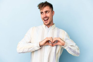 Mavi arka planda izole edilmiş genç beyaz adam gülümsüyor ve elleriyle kalp şeklini gösteriyor..