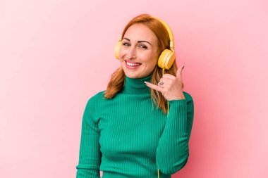 Pembe arka planda izole edilmiş müzik dinleyen genç beyaz kadın parmaklarıyla bir cep telefonu çağrısı hareketi gösteriyor..