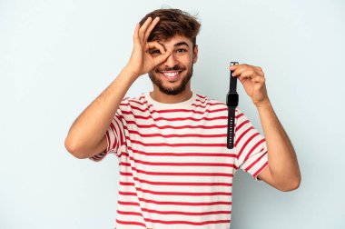 Young mixed race man holding a wrist watch isolated on blue background excited keeping ok gesture on eye.