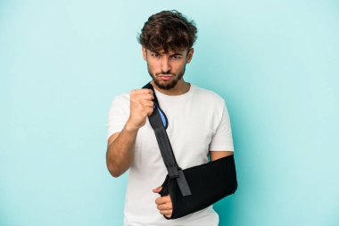 Young arab man with broken hand isolated on blue background showing fist to camera, aggressive facial expression.