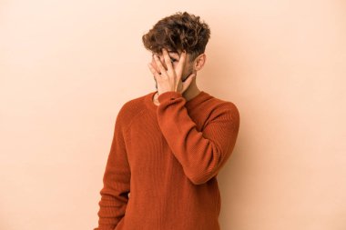 Young arab man isolated on beige background blink at the camera through fingers, embarrassed covering face.