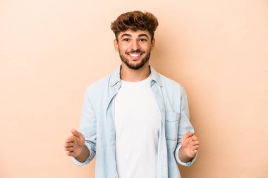 Young arab man isolated on beige background holding something with both hands, product presentation.