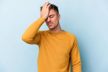 Kafkasyalı genç adam mavi arka planda izole edilmiş bir şekilde bir şeyi unutuyor. Alnına avuç içi ve gözlerini kapayarak vuruyor..