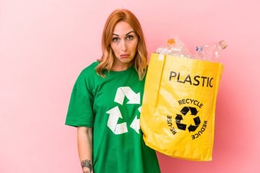 Young caucasian woman recycled plastic isolated on pink background shrugs shoulders and open eyes confused.