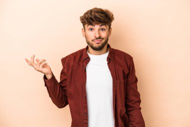 Young arab man isolated on beige background doubting and shrugging shoulders in questioning gesture.