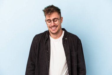 Mavi arka planda izole edilmiş genç beyaz adam gülüyor ve gözlerini kapıyor, rahatlamış ve mutlu hissediyor..