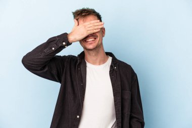 Mavi arka planda soyutlanmış genç beyaz bir adam elleriyle gözlerini kapatıyor, geniş bir gülümsemeyle bir sürpriz bekliyor..