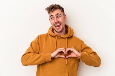 Beyaz arka planda izole edilmiş genç beyaz bir adam gülümsüyor ve elleriyle kalp şekli gösteriyor..