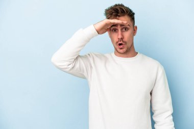 Kafkasyalı genç adam mavi arka planda izole edilmiş, uzaklara bakıyor ve elini alnına koyuyor..