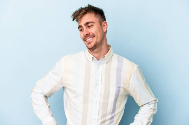 Kafkasyalı genç, mavi arka planda izole edilmiş. Kendine güvenen, kalçaları tutan biri..