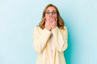 Mavi arka planda izole edilmiş genç beyaz kadın şok olmuş, ağzı ellerle kaplanmış, yeni bir şey keşfetmeye hevesli..