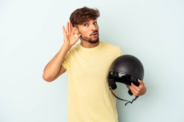 Young mixed race man holding motorcycle helmet isolated on blue background trying to listening a gossip.