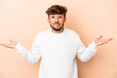 Young arab man isolated on beige background confused and doubtful shrugging shoulders to hold a copy space.