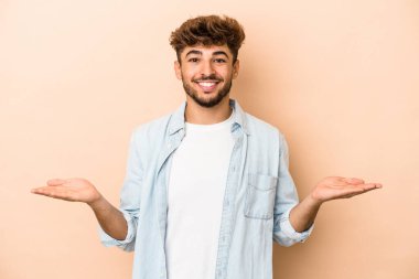 Young arab man isolated on beige background makes scale with arms, feels happy and confident.