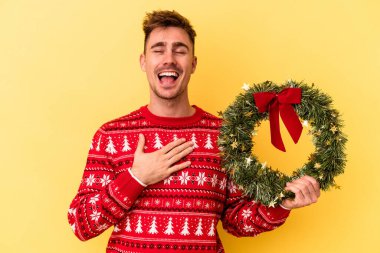 Sarı arka planda izole edilmiş bir çelenk tutan genç beyaz adam elini göğsünde tutarak kahkahalar atıyor..