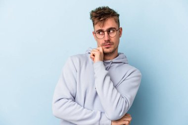 Kafkasyalı genç adam mavi arka planda izole, kafası karışmış, şüpheli ve güvensiz hissediyor..
