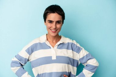 Kafkasyalı genç bir kadın mavi arka planda izole edilmiş. Kendine güvenen, ellerini kalçasında tutan..