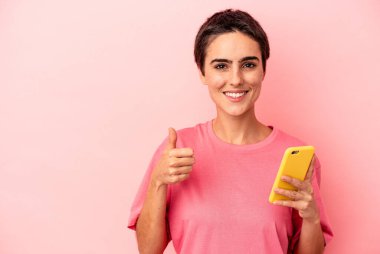 Elinde cep telefonu olan beyaz bir kadın pembe arka planda izole edilmiş gülümsüyor ve başparmağını kaldırıyor.