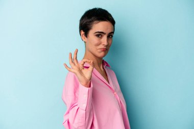 Kafkasyalı genç bir kadın mavi arka planda izole edilmiş bir şekilde bir göz kırpıyor ve elleriyle iyi bir jest yapıyor..