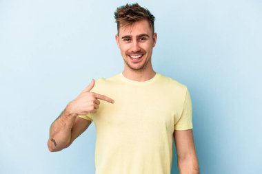 Mavi arka planda izole edilmiş genç beyaz adam bir gömlek fotokopi alanını işaret ediyor, gururlu ve kendinden emin.