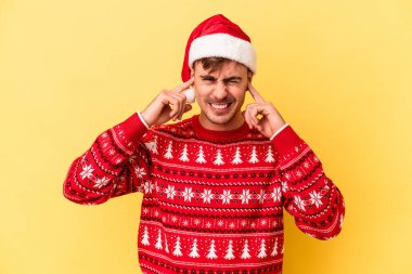Genç beyaz adam Noel 'i kutluyor. Sarı arka planda izole edilmiş. Kulakları elleriyle kapatıyor..