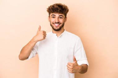 Young arab man isolated on beige background raising both thumbs up, smiling and confident.
