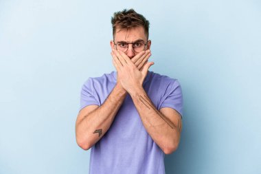 Mavi arka planda izole edilmiş genç beyaz adam elleriyle ağzını kapatırken şok oldu..