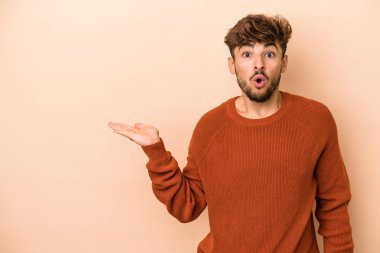 Young arab man isolated on beige background impressed holding copy space on palm.