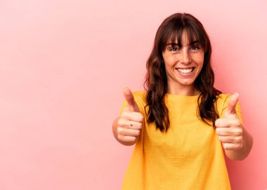 Arjantinli genç bir kadın pembe arka planda izole edilmiş gülümsüyor ve başparmağını kaldırıyor.