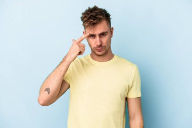 Mavi arka planda izole edilmiş genç beyaz bir adam şakağı parmağıyla işaret ediyor, düşünüyor ve bir göreve odaklanıyor..