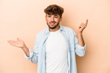 Young arab man isolated on beige background holding and showing a product on hand.