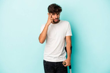 Young arab man with crutches isolated on blue background showing a disappointment gesture with forefinger.
